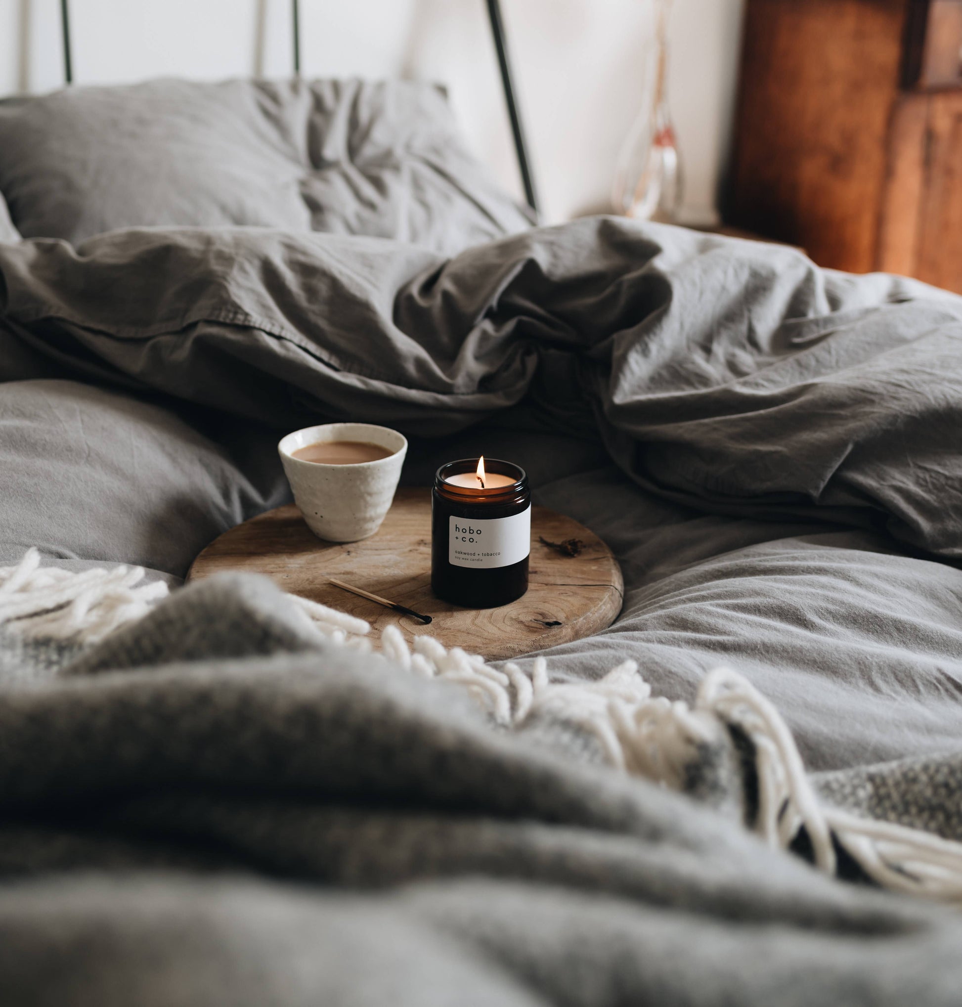 Candle and cup on a wooden tray on a bed with gray bedding